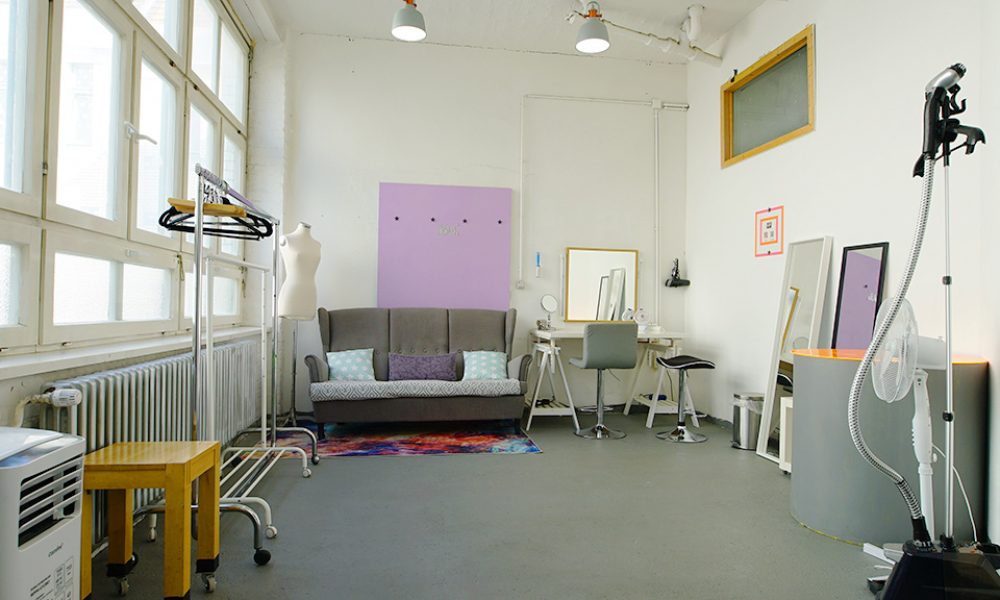 Heller Make-up-Raum im Allmost Mietstudio Berlin mit grauer Couch, Schminktisch, Spiegel, Kleiderständer und buntem Teppich für professionelle Film- und Fotoshootings