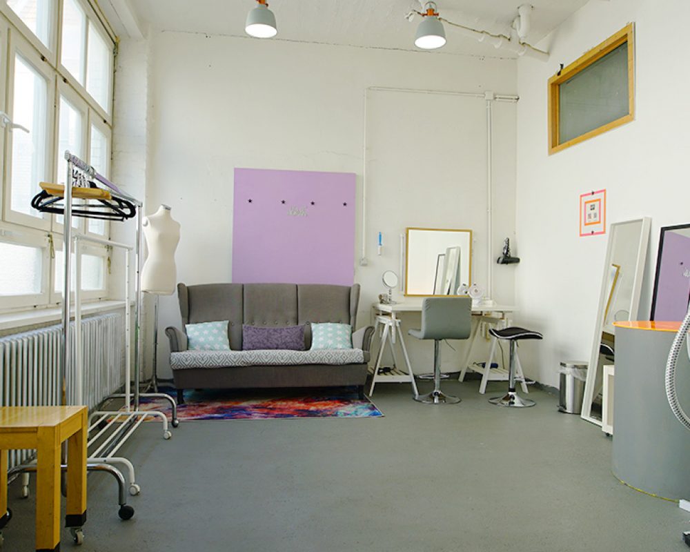Heller Make-up-Raum im Allmost Mietstudio Berlin mit grauer Couch, Schminktisch, Spiegel, Kleiderständer und buntem Teppich für professionelle Film- und Fotoshootings